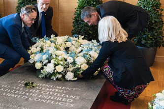 Drei Männer und eine Frau legen auf einem Grabstein mit Inschrift einen Blumenkranz nieder.