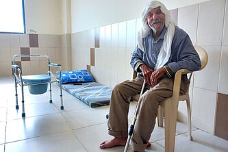 Moustafa al Sweid sitzt mit einem Gehstock lächelnd in einem kargen, aber sauberem Raum.