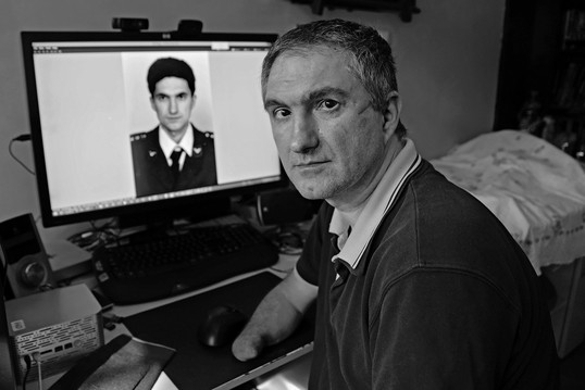 Branislav Kapetanović schaut ernst in die Kamera, hinter ihm ein Computerbildschirm mit einem Bild von ihm als junger Mann.