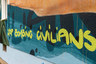 Graffiti der Ausstellungsreihe „Stop Bombing Civilians“ von Handicap International mit blauem Hintergrund und der Aufschrift "Stop Bombing Civilians" in Gelb 
