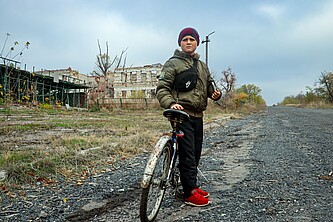 Nazar lehnt an sein rostiges Fahrrad. Im Hintergrund seine zerbombte Schule.