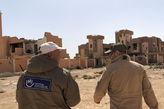 Zwei Männer gehen auf eine Reihe bombardierter Häuser zu. Die Gegend ist wüstenähnlich.